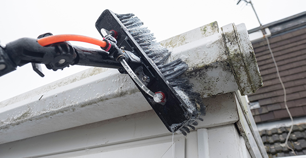 a person using a brush attachment on a pressure washer cleaning the exterior of a gutter